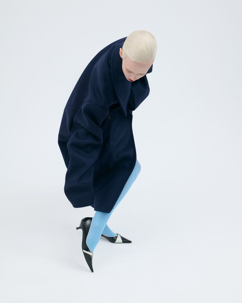 A woman with a short blonde haircut wears a dark blue coat and light blue tights with black heels against a bright white background.