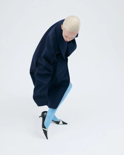 A woman with a short blonde haircut wears a dark blue coat and light blue tights with black heels against a bright white background.
