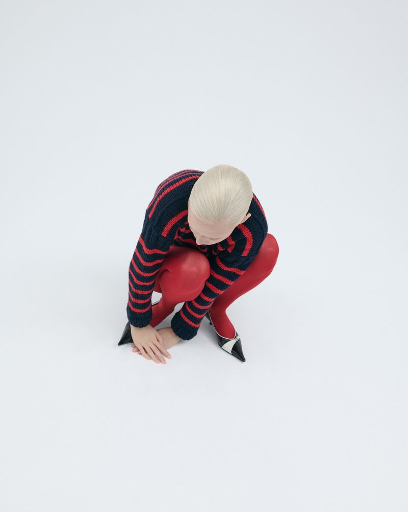 Blonde woman in blue and red sweater and red tights crouching on white seamless background looking at her hands and wearing high heels.