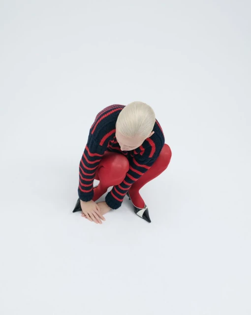 Blonde woman in blue and red sweater and red tights crouching on white seamless background looking at her hands and wearing high heels.
