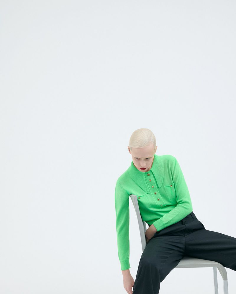 An adult female with light blonde hair, dressed in a neon green long-sleeve shirt and black pants, sits on a gray chair in a photo studio.