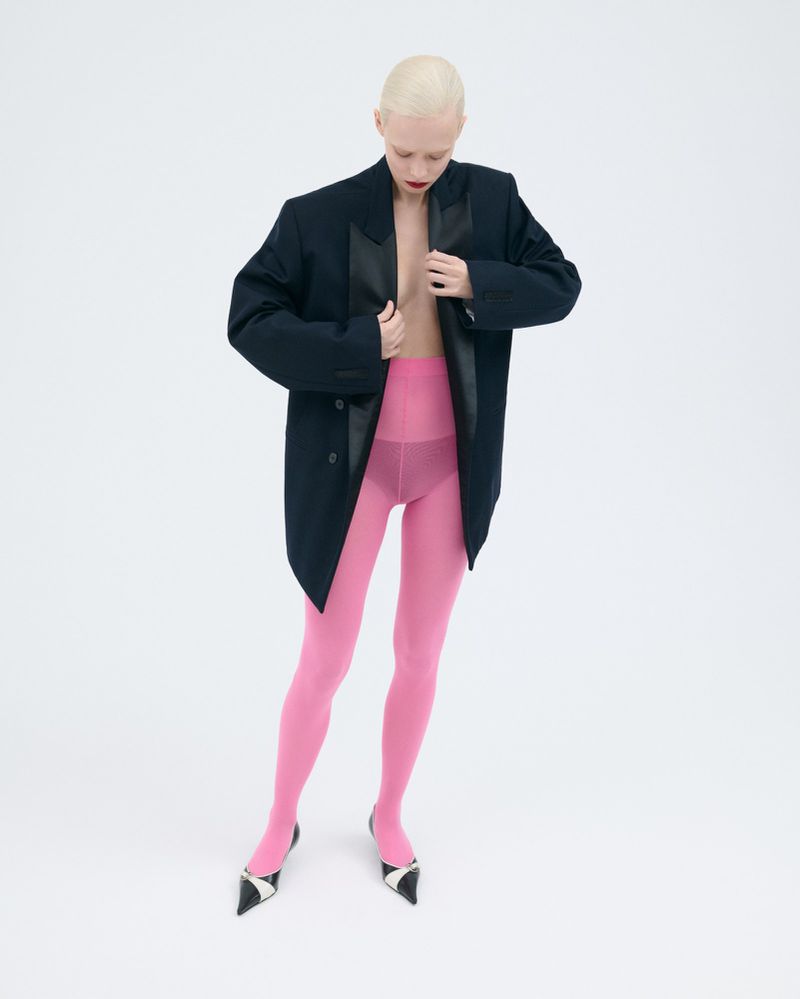 Full-length studio shot of a woman in a dark blazer, pink tights, and black and white heels posing against a white backdrop.