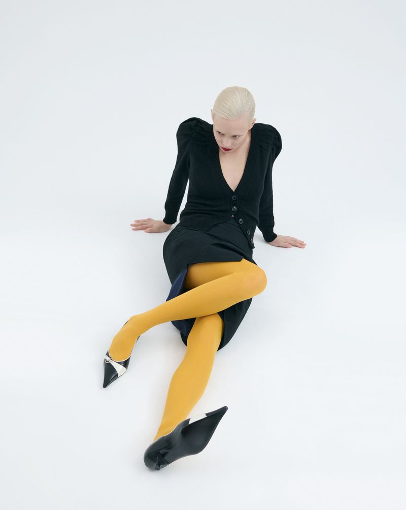 Woman sitting on a white floor in a dark skirt and sweater with yellow tights and black pointed shoes in a fashion style pose.