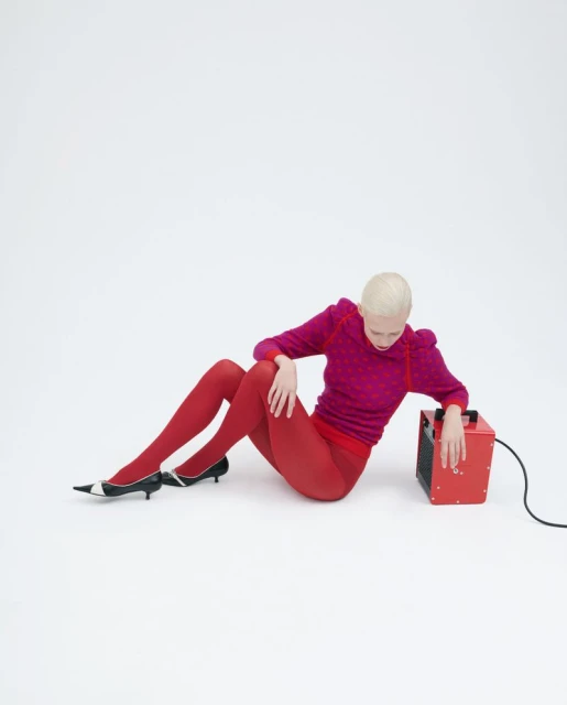 An adult woman poses seated on the ground next to a red space heater in a studio environment with white background and seamless.