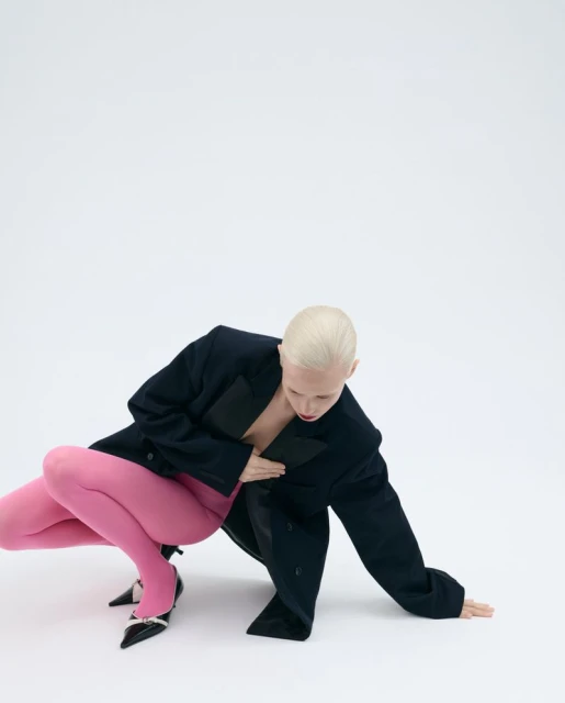 Woman in a black jacket and pink tights squats indoors on a white background, creating a high-fashion pose for a fashion editorial.