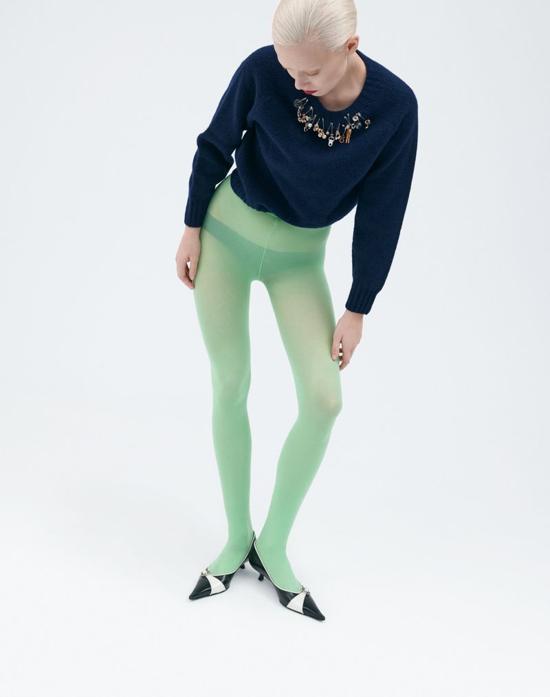 Studio shot of a woman wearing a navy blue sweater with embellishments, light green tights, and black and white heels against a white backdrop.