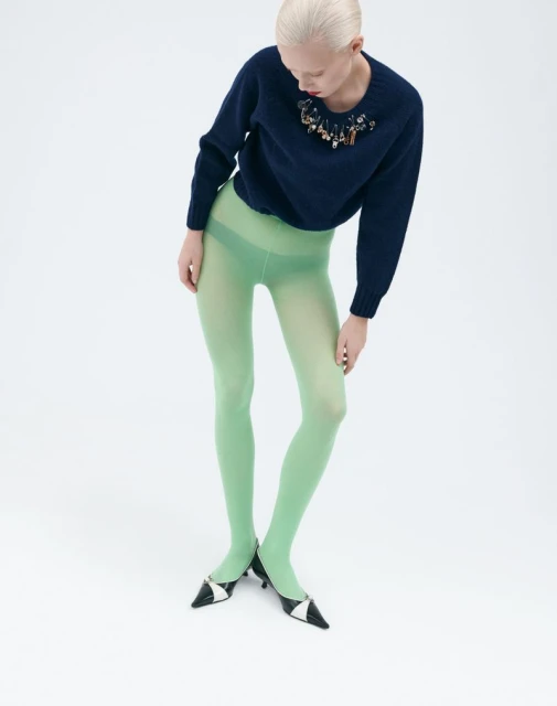 Studio shot of a woman wearing a navy blue sweater with embellishments, light green tights, and black and white heels against a white backdrop.