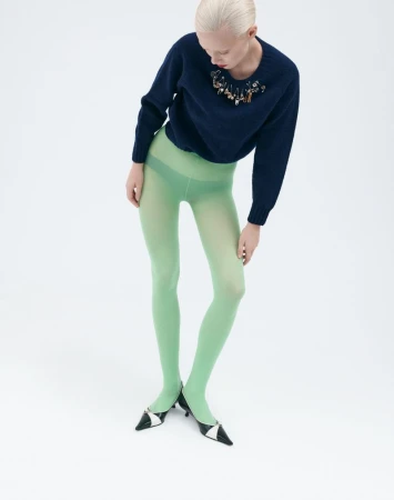 Studio shot of a woman wearing a navy blue sweater with embellishments, light green tights, and black and white heels against a white backdrop.