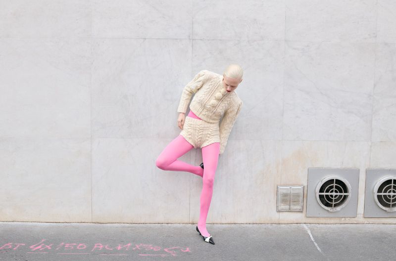 A woman with short blonde hair and a knitted outfit poses against a gray wall, wearing pink stockings and black and white shoes.