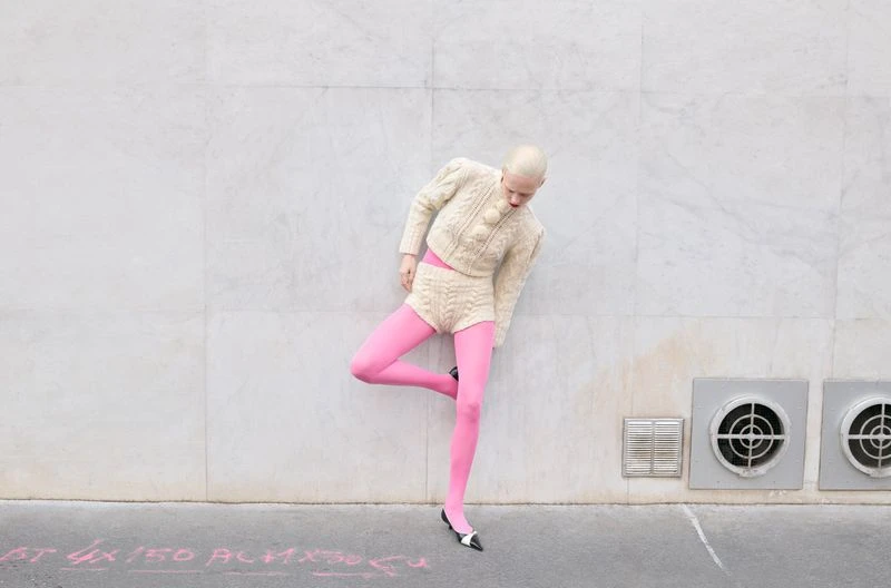 A woman with short blonde hair and a knitted outfit poses against a gray wall, wearing pink stockings and black and white shoes.