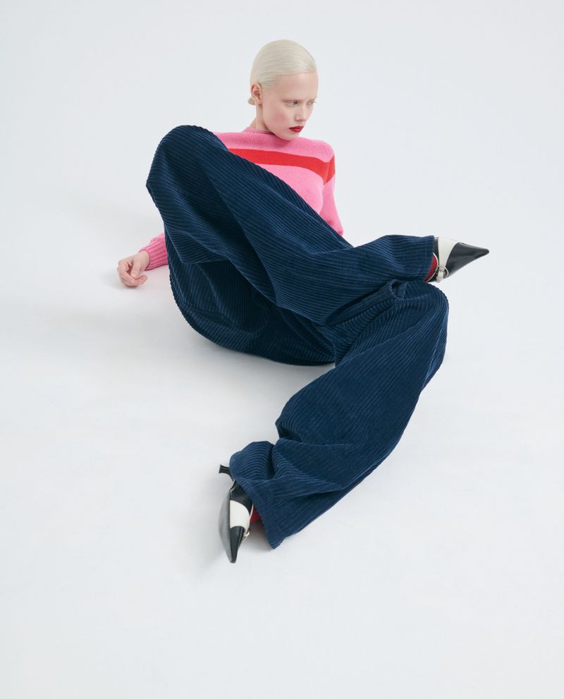 Fashion shot of a woman with short platinum hair wearing a pink and red striped sweater and navy corduroy pants on a white background.
