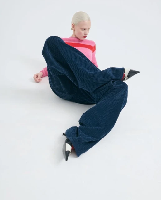 Fashion shot of a woman with short platinum hair wearing a pink and red striped sweater and navy corduroy pants on a white background.