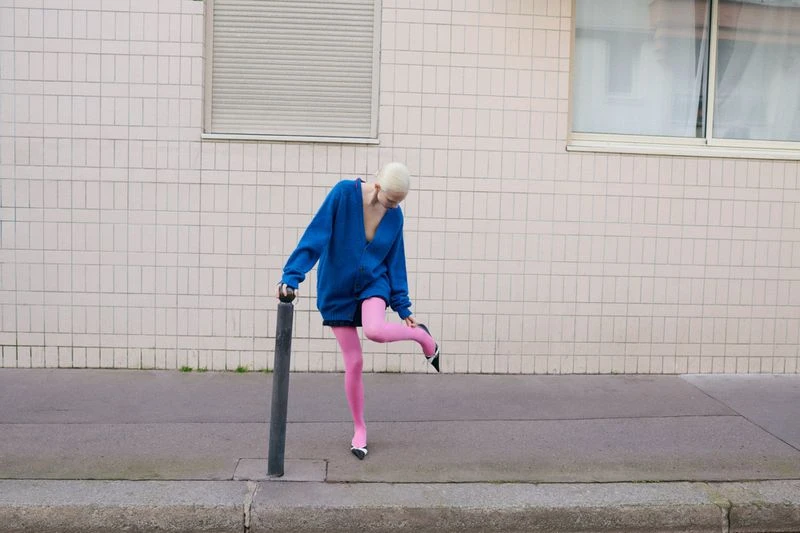 Woman in oversized blue sweater and pink tights adjusts black and white high heel on city sidewalk, leaning against a black pole.