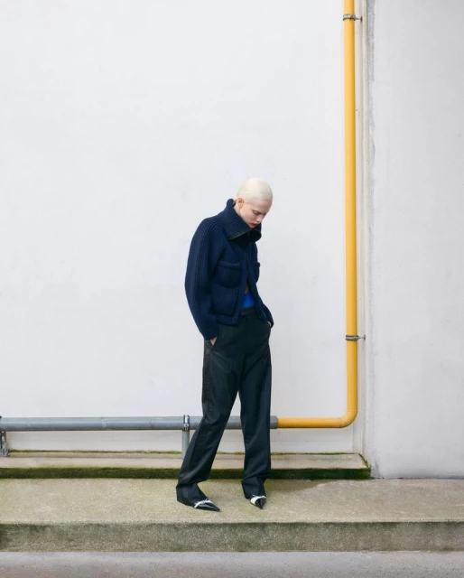 A fair-skinned person with shaved platinum hair poses in a navy jacket and black pants against a white wall and concrete steps outside.