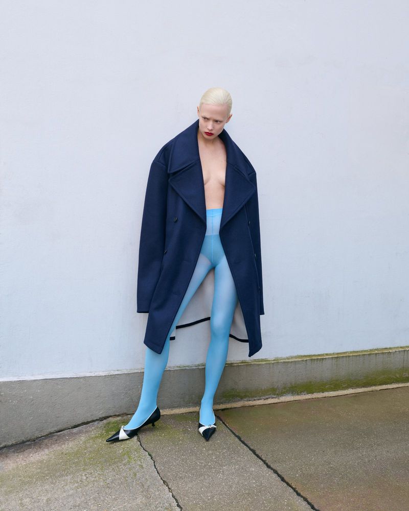 A fair-skinned person with a shaved head, wearing a navy coat and light blue tights, poses in an outdoor fashion setting against a white wall.