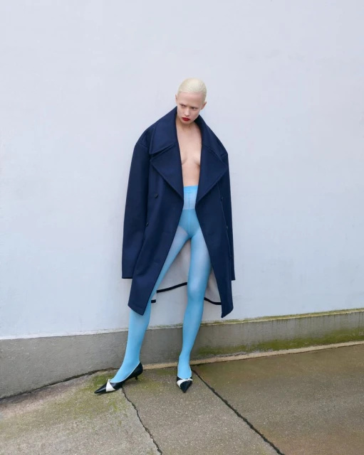A fair-skinned person with a shaved head, wearing a navy coat and light blue tights, poses in an outdoor fashion setting against a white wall.