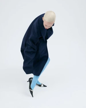 High-angle view of a blonde model wearing an oversized navy coat, light blue tights, and black pointed heels on a white studio background.