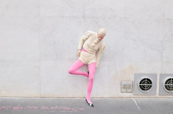 Model with a blonde buzz cut wearing a cream cable-knit set and bright pink tights posing against a light grey stone wall.