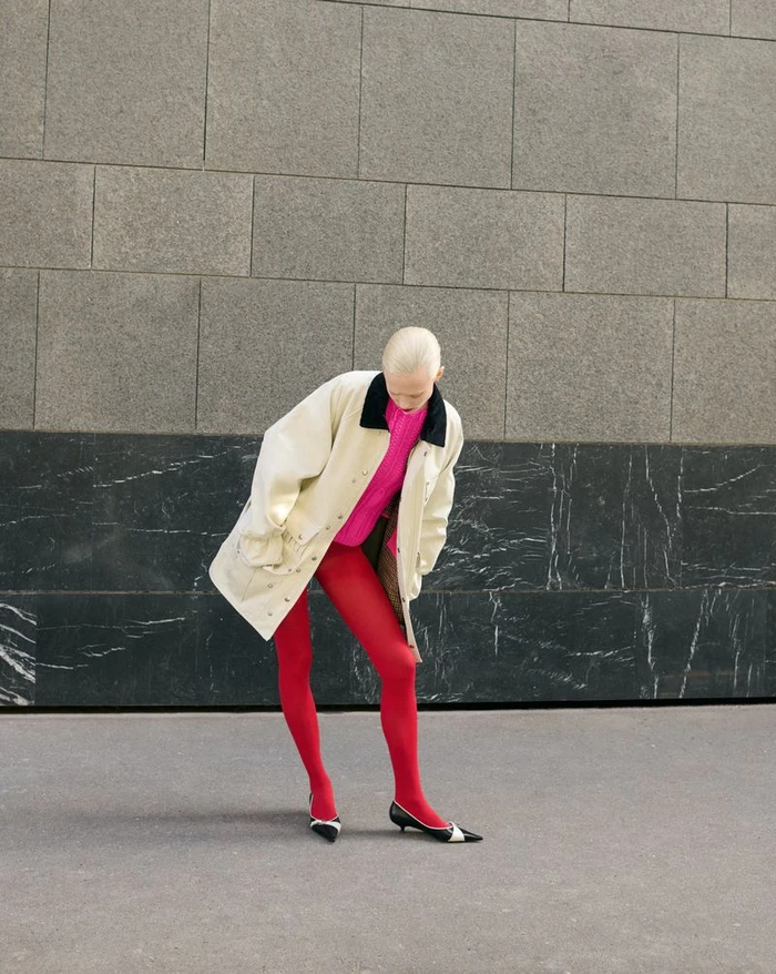 Model in an oversized cream coat, bright pink top, red tights, and pointed kitten heels posing against an outdoor stone wall.