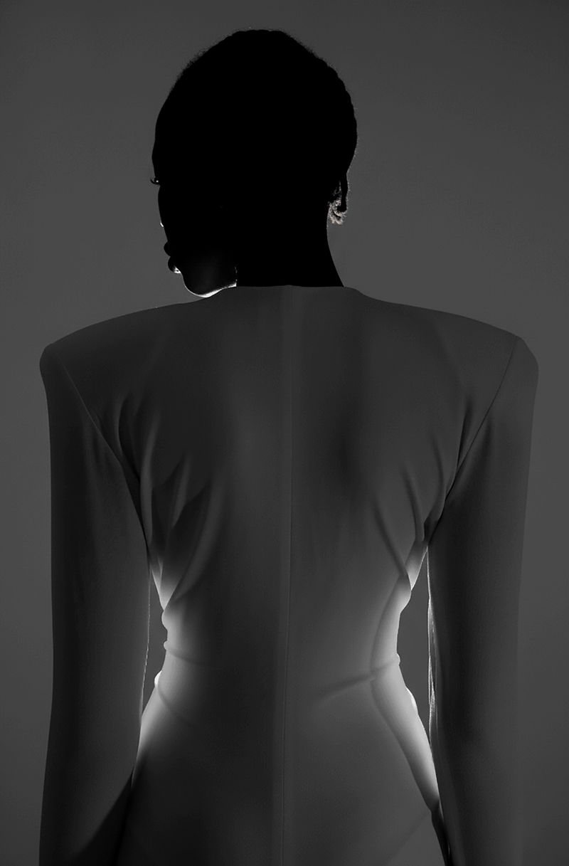Monochrome portrait of a slender, dark-skinned woman wearing a white, long-sleeved dress; shot in a photo studio against a gray background.