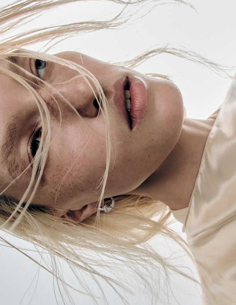 Close-up portrait of Liza Ostanina with windblown blonde hair wearing a cream satin top and silver earring against a pale background.