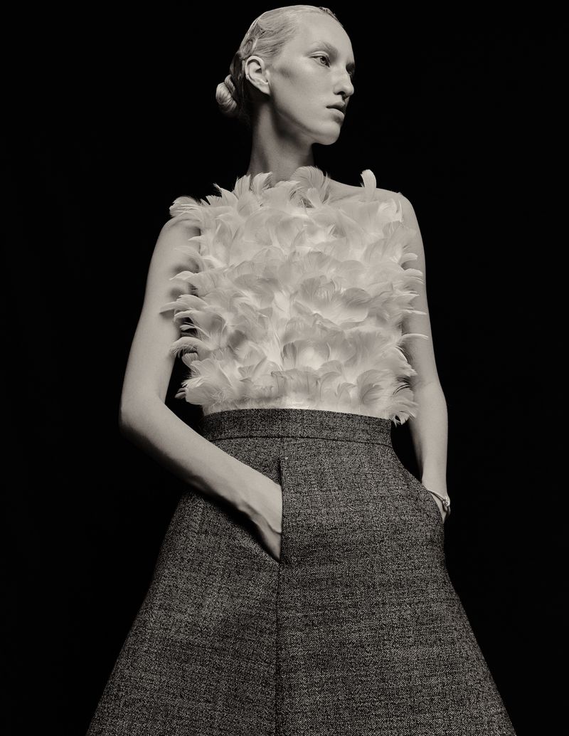 Black and white fashion shot of a woman in a feather top and skirt, posing with her hand in her pocket, against a black background.