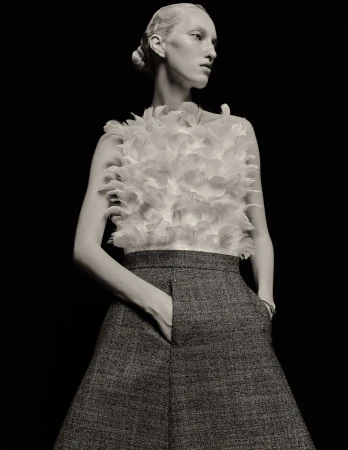 Black and white fashion shot of a woman in a feather top and skirt, posing with her hand in her pocket, against a black background.