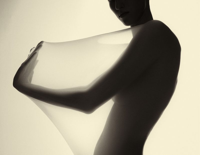 Artistic studio shot of a figure draped with sheer white fabric, set against a soft background with muted sepia tones.