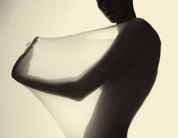 Artistic studio shot of a figure draped with sheer white fabric, set against a soft background with muted sepia tones.