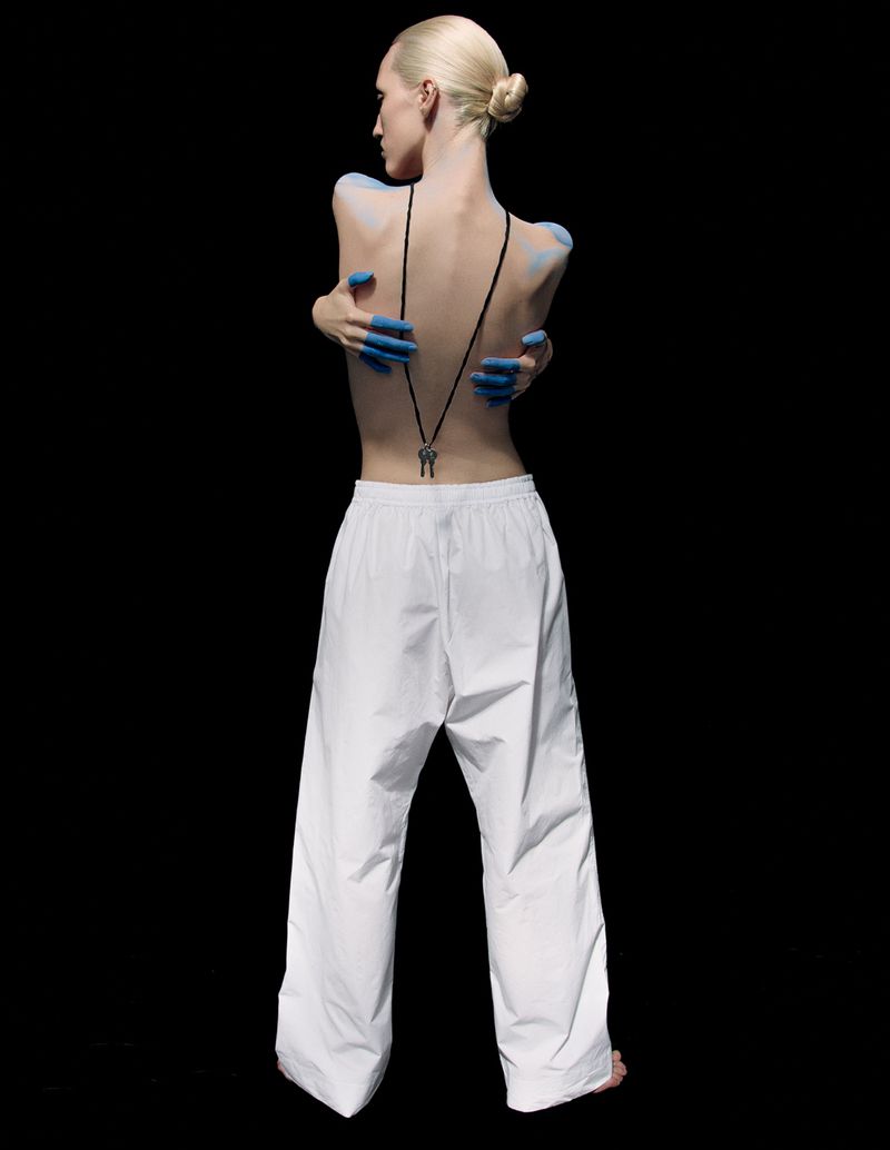 Back view of a fair-skinned adult female with light blonde hair posing in white pants, blue painted hands and pendant necklace on black.