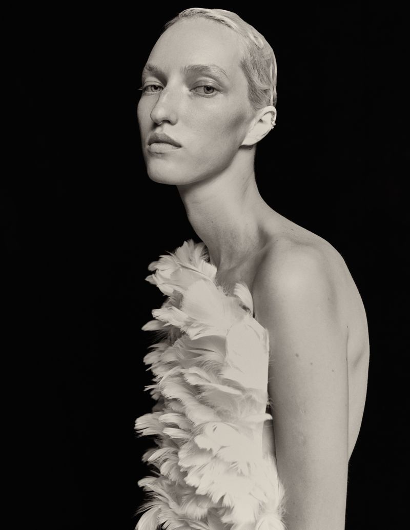 Monochrome portrait of a slender adult model with short blonde hair and a feathered garment against a solid black backdrop. 