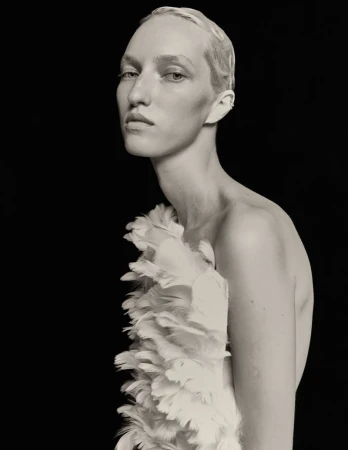 Monochrome portrait of a slender adult model with short blonde hair and a feathered garment against a solid black backdrop. 