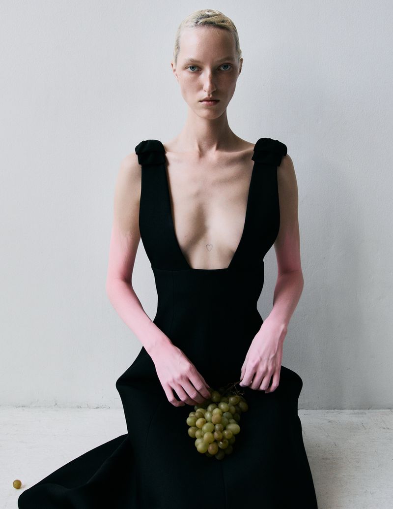 Woman in black dress holds grapes against a white background, fashion editorial, studio lighting, high fashion, elegant style.