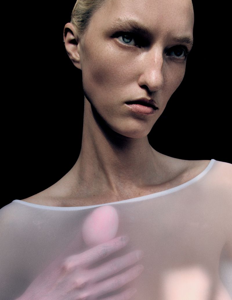 Portrait of a fair-skinned woman with blonde hair, wearing a sheer white top, against a stark black background in a studio setting.