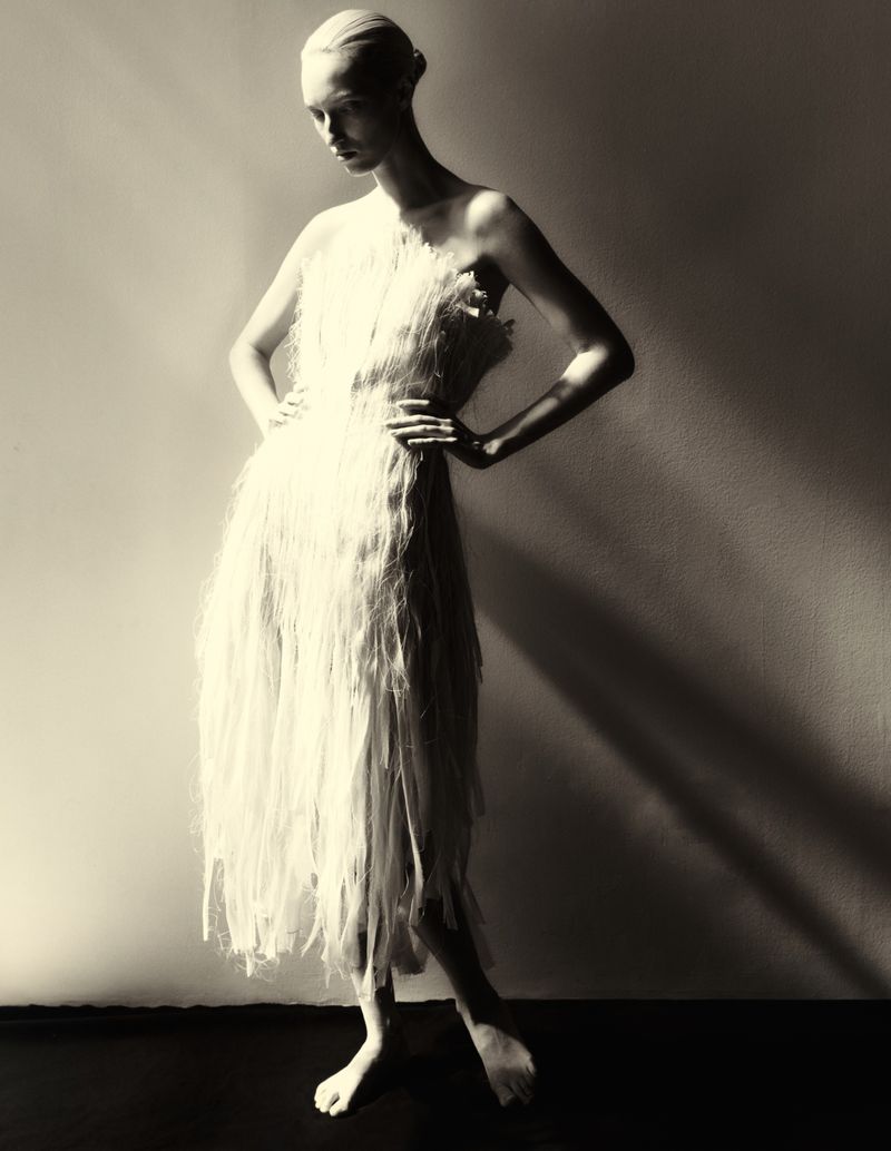 Full-length portrait of a female fashion model in an off-white shredded fabric dress, posing indoors with hands on hips.