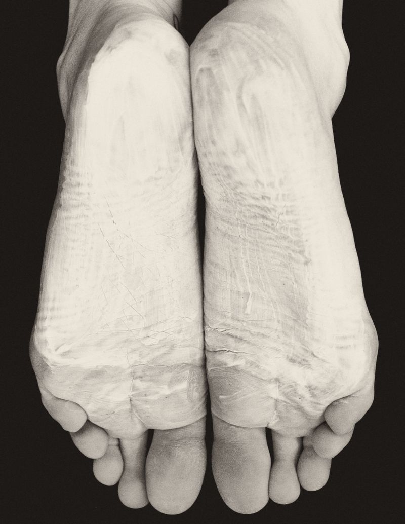 Close-up black and white image of two adult feet side by side with a light-colored cream or other material applied to them in a studio.