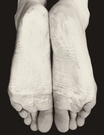 Close-up black and white image of two adult feet side by side with a light-colored cream or other material applied to them in a studio.
