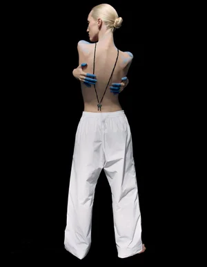 Model Liza Ostanina viewed from behind with blue-painted hands on her bare back wearing wide white pants against a black background.