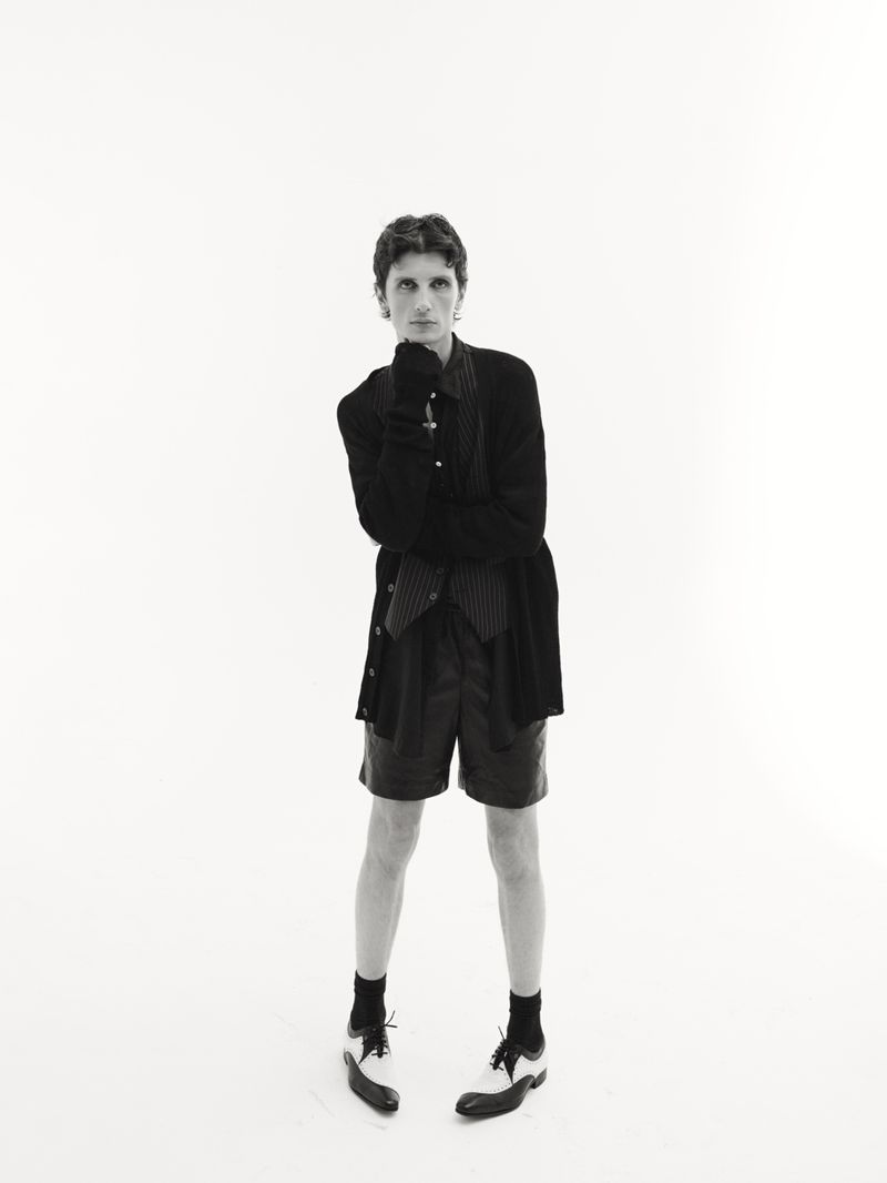 Black and white studio portrait of a young adult posing in pinstripe vest and shorts with cardigan, socks, and two-tone wingtip shoes.