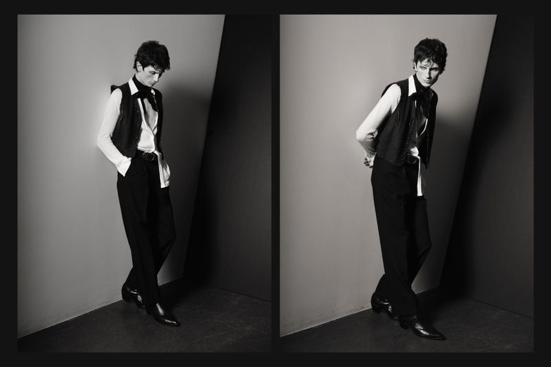 Black-and-white fashion shot of a young adult man in a vest, dress shirt, and trousers leaning against a corner in a studio environment.