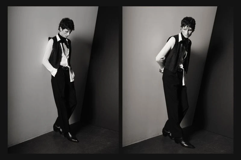 Black-and-white fashion shot of a young adult man in a vest, dress shirt, and trousers leaning against a corner in a studio environment.
