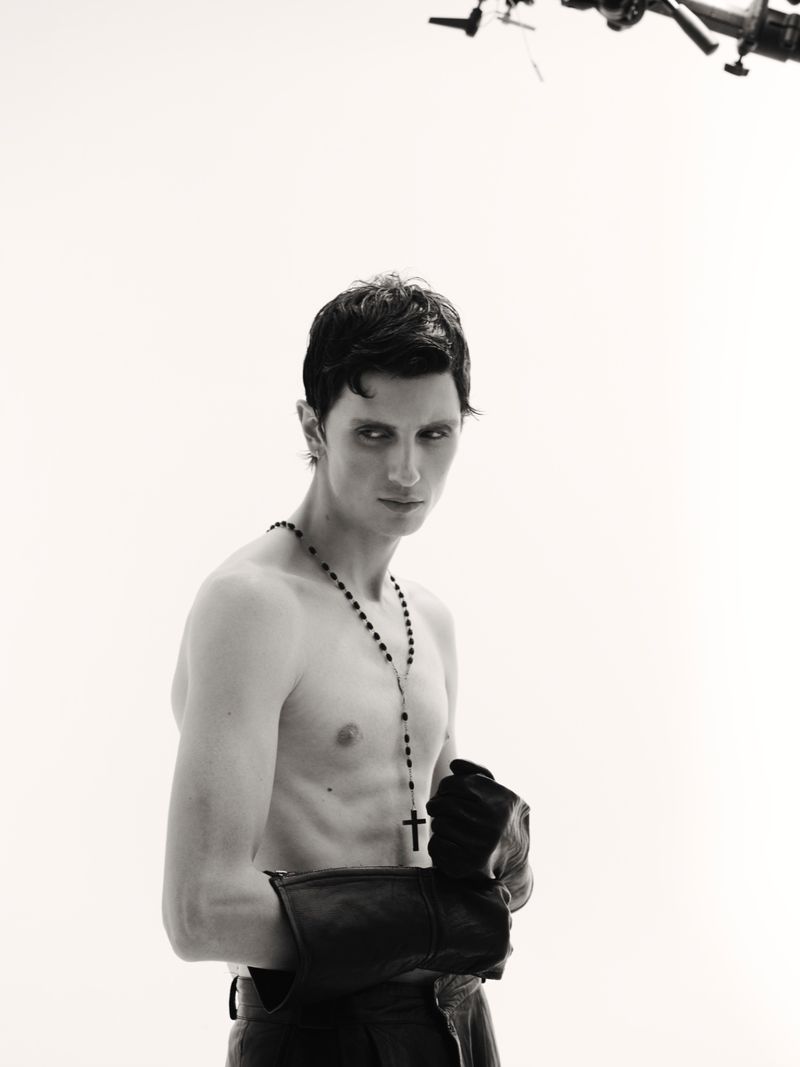 Monochrome portrait of a shirtless adult male with dark hair, wearing leather gloves and a rosary necklace, against a bright backdrop.