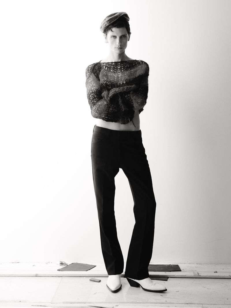 Monochrome portrait of a slender man in a knit top and dark pants with white boots, standing against a pale, neutral backdrop in studio.