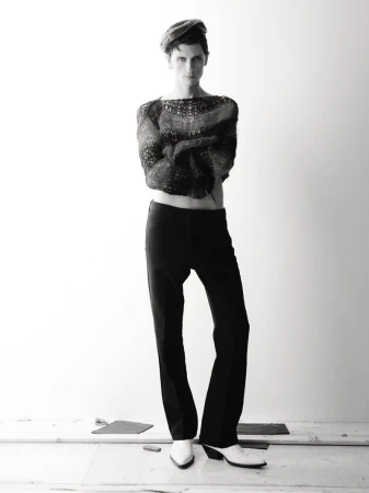 Monochrome portrait of a slender man in a knit top and dark pants with white boots, standing against a pale, neutral backdrop in studio.