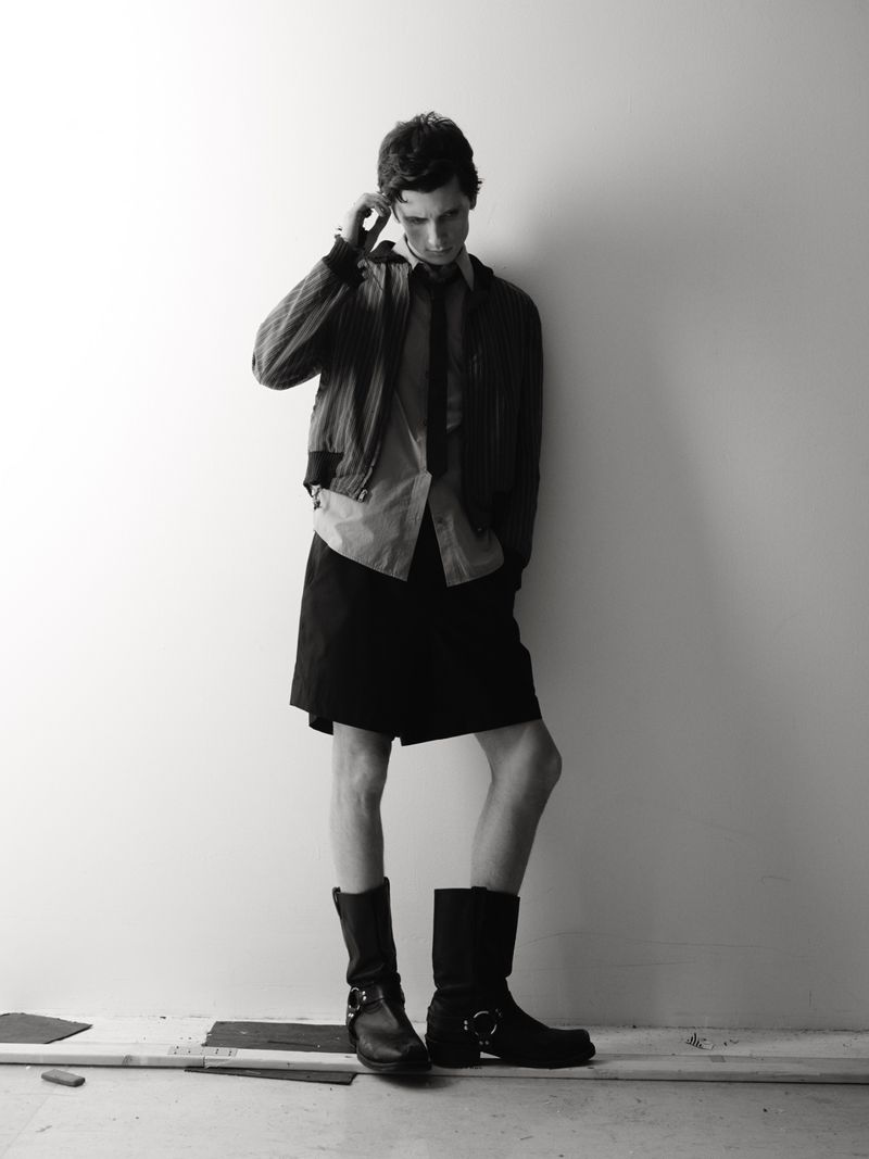 Monochrome fashion portrait of a young adult man in jacket, skirt, and boots posing against a white wall in a studio setting.