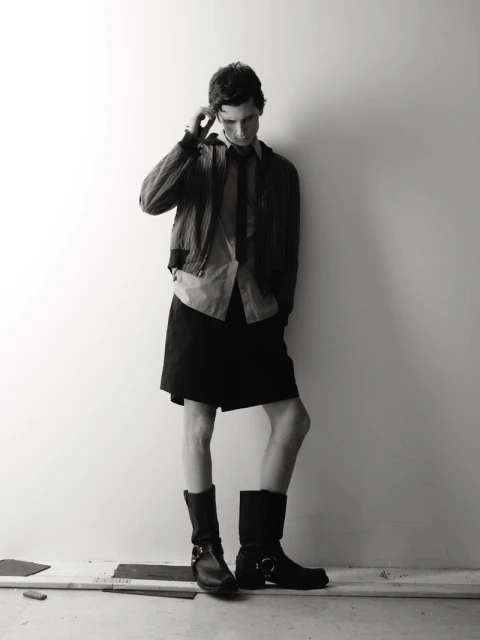 Monochrome fashion portrait of a young adult man in jacket, skirt, and boots posing against a white wall in a studio setting.