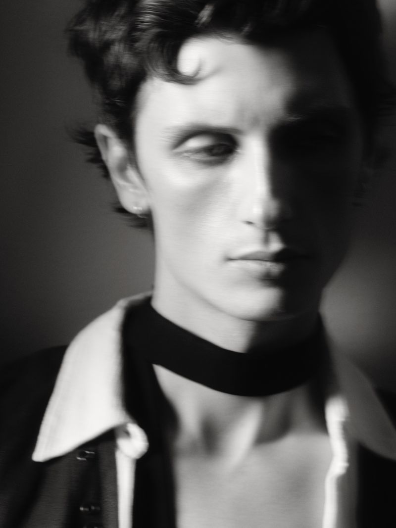 Portrait of a dark-haired adult male in black and white, wearing a blazer, white collared shirt, and black neck band. Soft focus.
