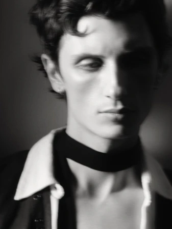 Portrait of a dark-haired adult male in black and white, wearing a blazer, white collared shirt, and black neck band. Soft focus.