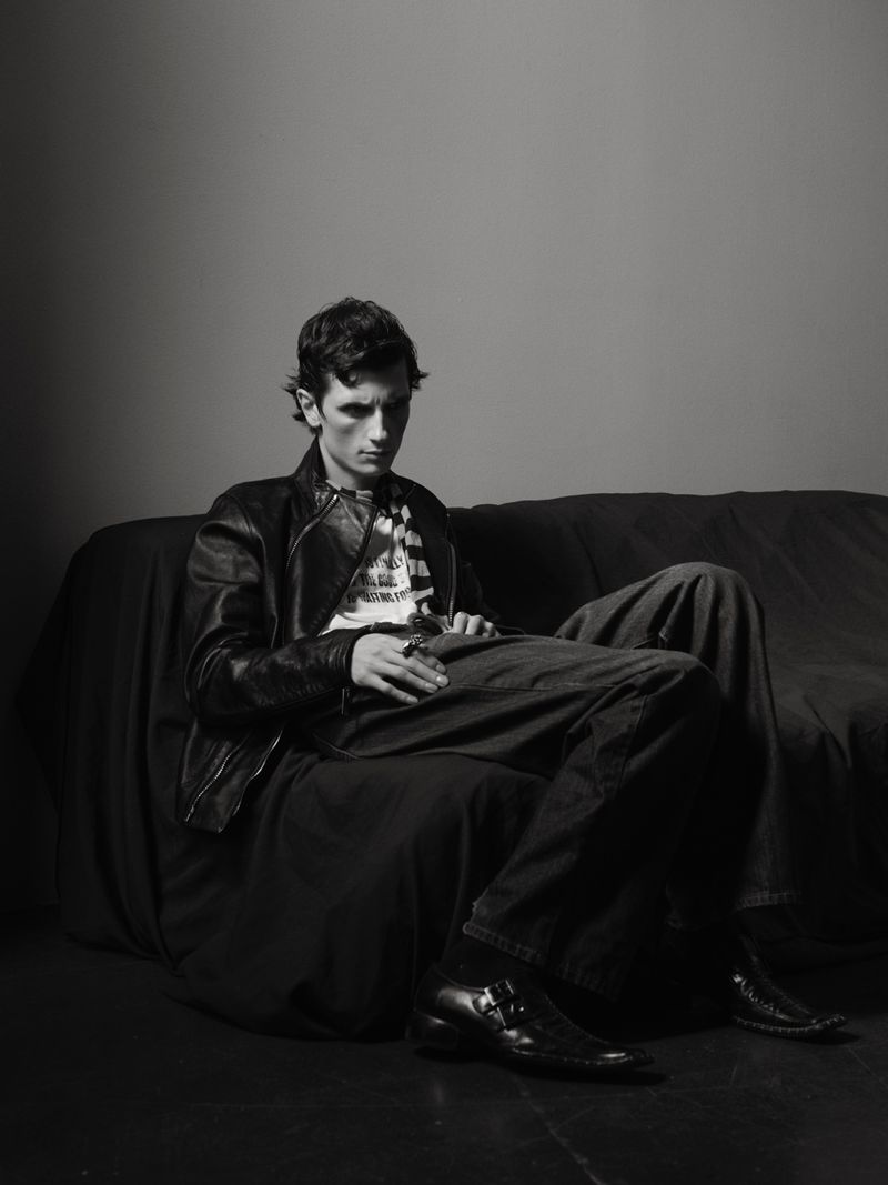 Young man with dark hair wearing a t-shirt, leather jacket, jeans and black boots sitting on a couch in a photo studio.