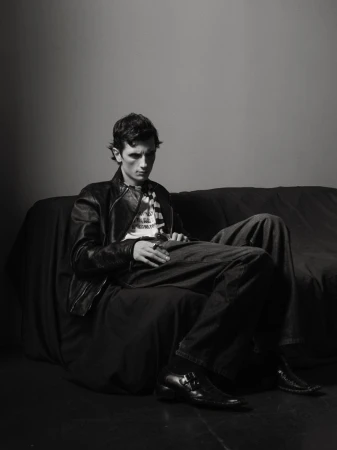 Young man with dark hair wearing a t-shirt, leather jacket, jeans and black boots sitting on a couch in a photo studio.